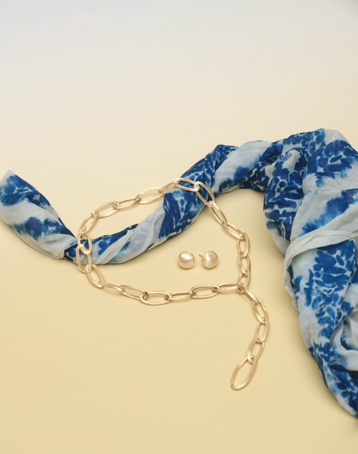A sleek gold chain necklace and complimentary stud earrings displayed on a yellow backdrop against a vibrant blue and white scarf evocative of ocean waves. A sleek gold chain necklace and complimentary stud earrings displayed on a yellow backdrop against a vibrant blue and white scarf evocative of ocean waves.