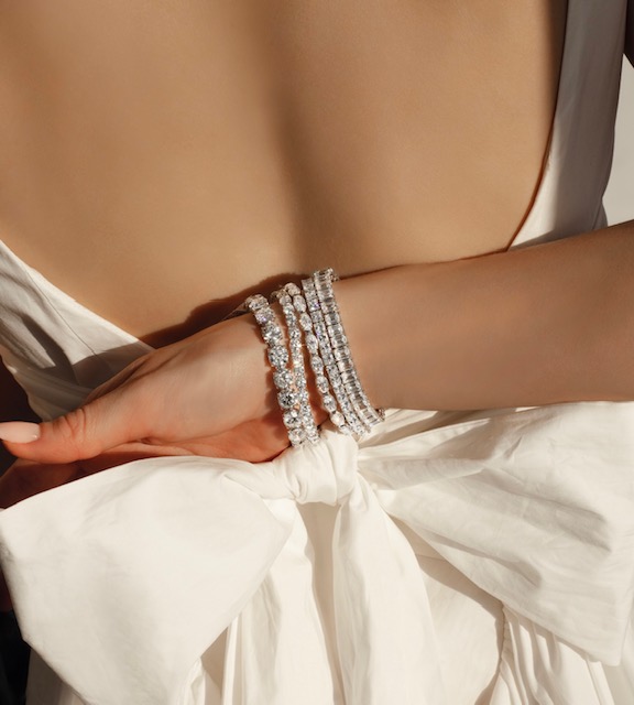 A close-up of an elegantly dressed woman’s hand behind her back, adorned with multiple stunning tennis bracelets. A close-up of an elegantly dressed woman’s hand behind her back, adorned with multiple stunning tennis bracelets.